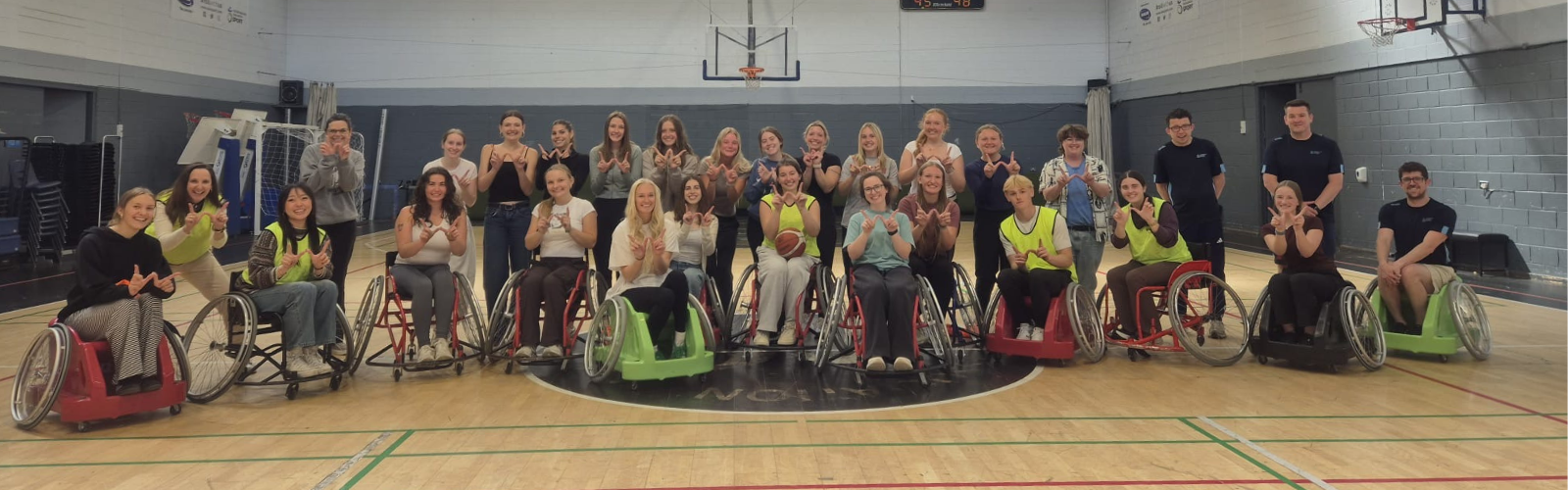 A group of students in a gym, some standing and some in wheelchairs, make the shape of a W with their fingers to represent UW-Madison.